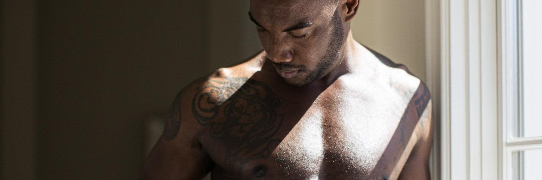 Man with chest tattoos standing by window in natural light, featured by JIMMYJANE luxury wellness.