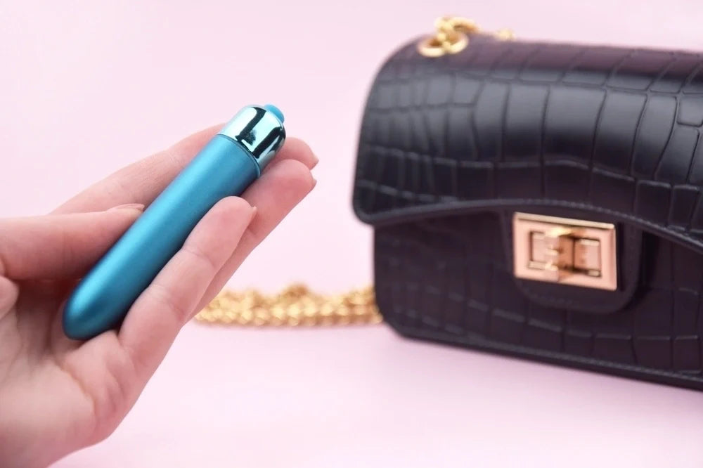 A hand holding a blue bullet vibe in front of a black patent leather purse.