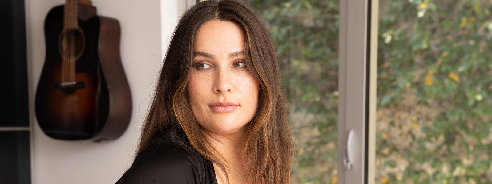 Woman with long brown hair in natural light indoors, acoustic guitar on wall, JIMMYJANE lifestyle