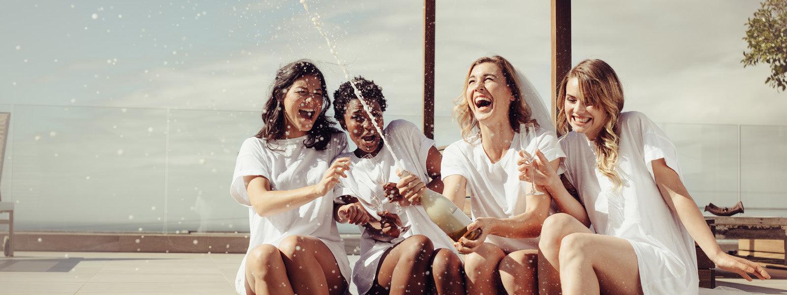 Four women in white shirts celebrating outdoors with champagne, smiling and laughing, luxury lifestyle