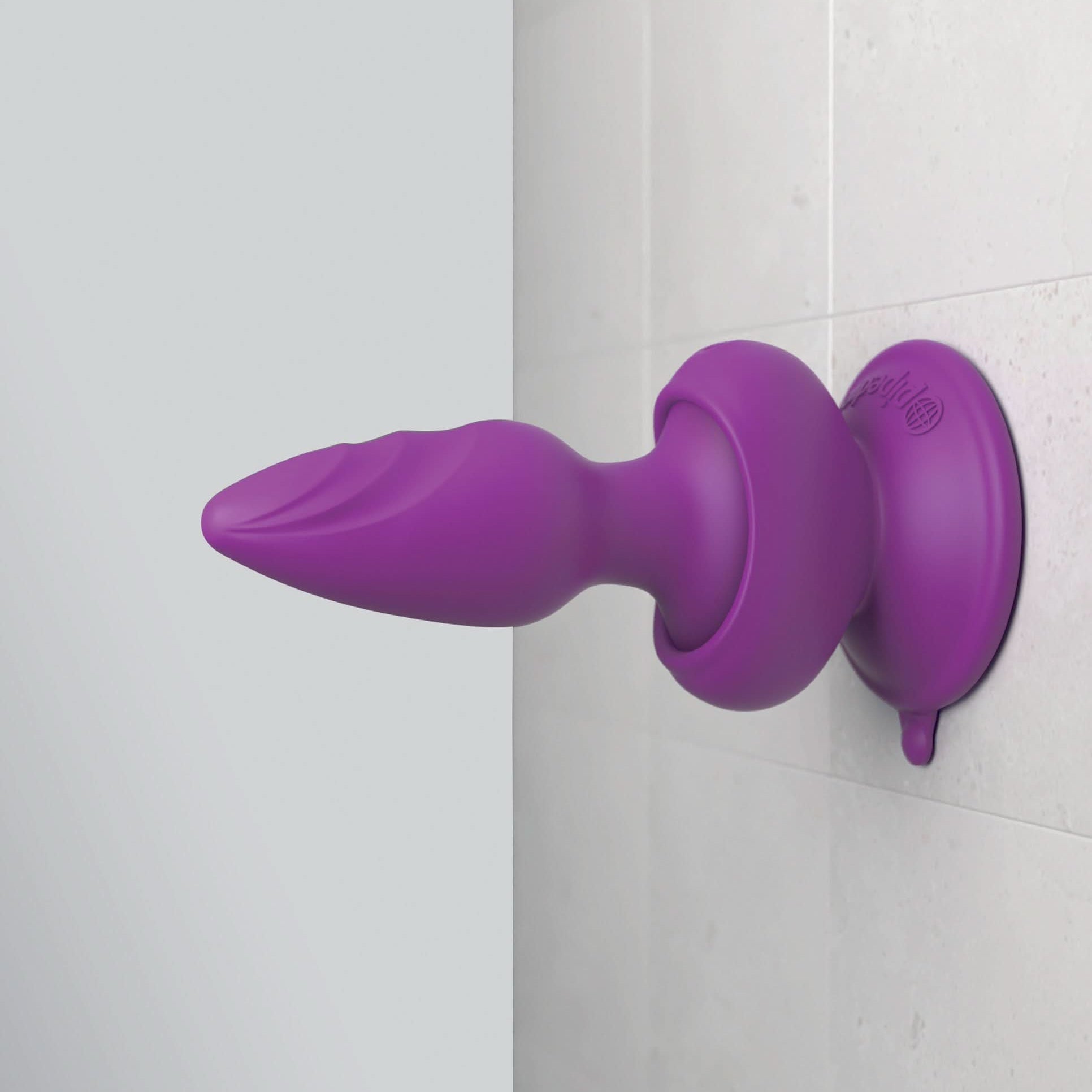 A Pipedream® Wall Banger Plug by 3Some, featuring a sturdy suction cup base, is attached to a tiled bathroom wall for hands-free pleasure.