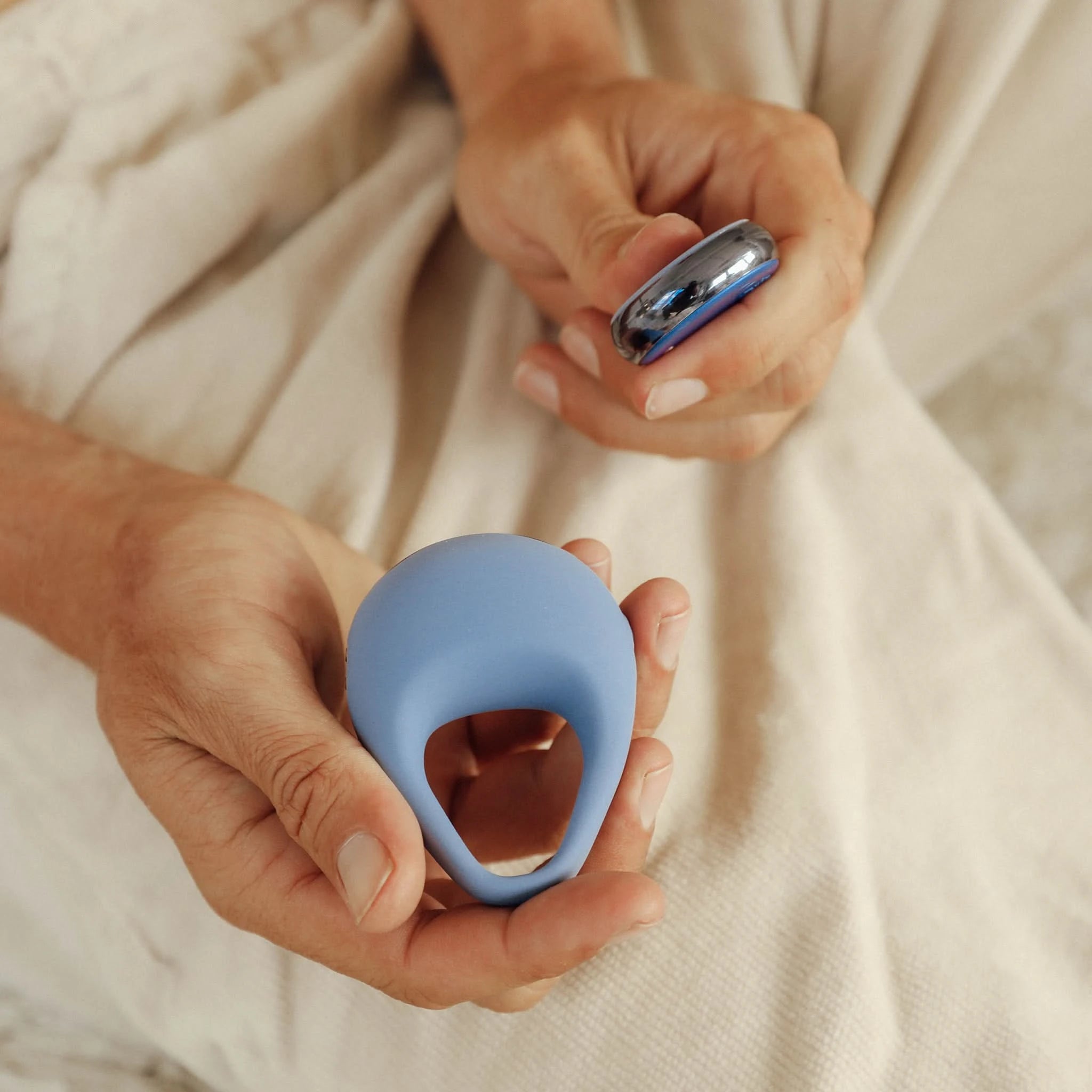 A person holds the JIMMYJANE® Kore™ with Remote, a blue vibrating silicone c-ring and its small remote control, while seated on light fabric.