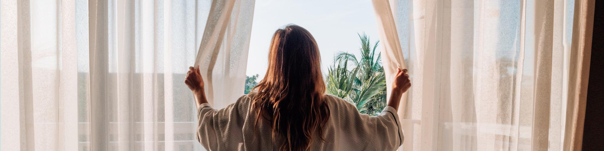 Woman in robe opening curtains to morning sunlight and palm trees, wellness self-care lifestyle