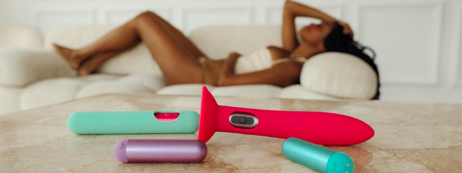 Luxury adult toys in vibrant colors on table, woman in lingerie lounging in background, JIMMYJANE