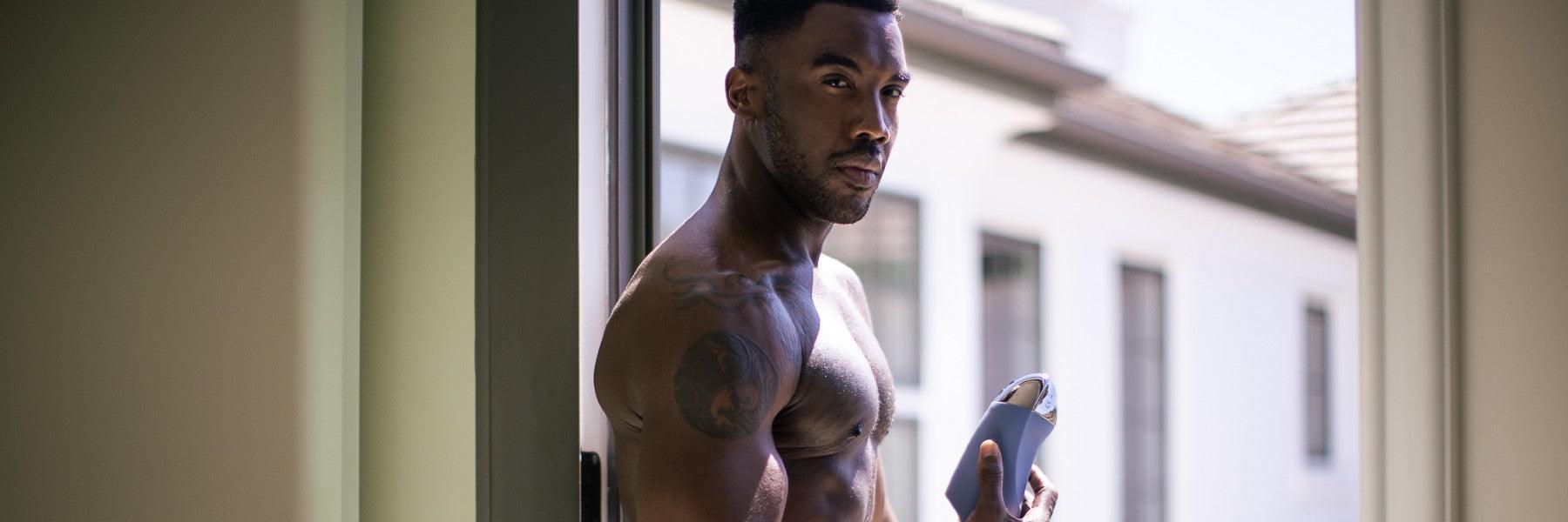 Fit man with tattoos holding sleek device by window, featuring JIMMYJANE luxury wellness product
