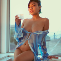 Young asian women wearing a sheer blue top draped over nude body sitting in front of an open window holding a lit afterglow vanilla sandalwood scented massage oil candle #vanilla sandalwood